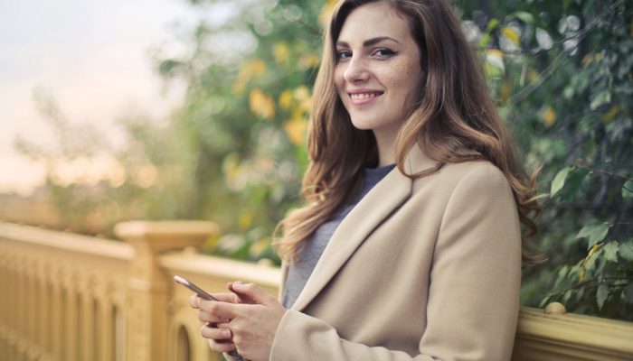 Selective Focus Photo of Smiling Woman in Brown Coat Holding Smartphone While Leaning on stone Railing